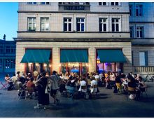 Straßenszene mit einem Café vor einem mehrstöckigen Gebäude mit weiß gefliester Fassade und großen Fenstern, geschmückt mit drei türkisfarbenen Markisen. Draußen sitzen zahlreiche Menschen an Tischen auf einem gepflasterten Platz, erleuchtet von warmem Licht aus dem Innenbereich.