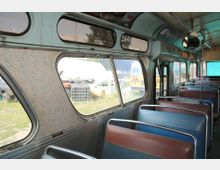 Innenraum eines alten Busses mit braunen und blauen Sitzbänken und großen Fenstern. Durch die Fenster ist eine Außenansicht mit geparkten Fahrzeugen und Gras zu erkennen.