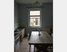 Kleine Küche mit einfacher Einrichtung, einem Holzesstisch in der Mitte und einem Fenster, durch das der Blick auf ein Fachwerkgebäude fällt. Links befinden sich ein Herd, eine Spüle und geflieste Wände; rechts steht ein Heizkörper.