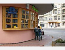 Straßenszene mit einem rosa Gebäude, das eine abgerundete Ecke und große Holzfenster besitzt, hinter denen Leuchtschilder sichtbar sind. Davor stehen gestapelte Stühle und Blumentöpfe, im Hintergrund sind Altbauten und Geschäfte an einer belebten Straße zu sehen.