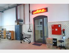 Raum mit weiß gefliesten Wänden und Industriestil, sichtbar sind ein Glastür-Eingang mit der Aufschrift „VARBLUT“, ein leuchtendes Neon-Schild darüber, Metallspinde, Regale mit Aktenordnern, ein Einkaufswagen und ein roter Erste-Hilfe-Schrank an der Wand.