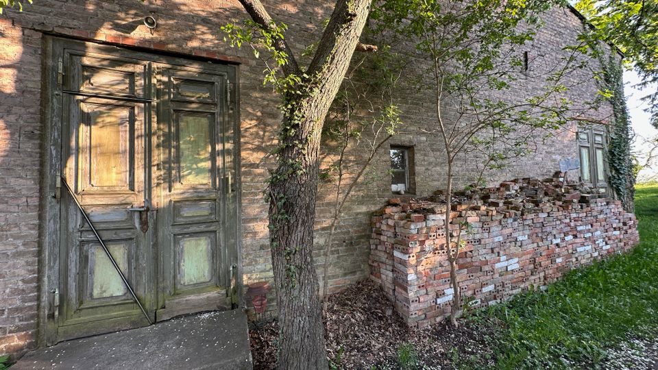 Außenansicht eines alten Backsteingebäudes mit verwitterter, grün gestrichener Holztür und kleiner Betontreppe. Daneben stehen ein Baum und ein hoher Stapel Ziegelsteine an der Wand, umgeben von Gras und niedrigem Bewuchs.