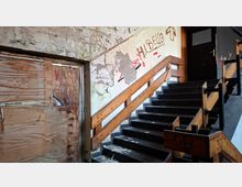 Verlassenes Treppenhaus in einem Gebäude mit dunklen Stufen und Holzgeländern, die zu einer geschlossenen Tür im oberen Stockwerk führen. Abblätternder Putz, Graffiti an der Wand und eine mit Sperrholz vernagelte Öffnung prägen den heruntergekommenen Innenraum.