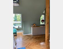 Ein Kinderzimmer mit grün gestrichenen Wänden, einem Fischgrät-Holzboden und einem großen Fenster, durch das Bäume sichtbar sind. Im Raum stehen eine Kommode, ein kleiner Spielzeugladen sowie ein blauer Sessel, und ein Holzbalken ist sichtbar.