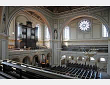 Innenraum einer Kirche mit hohen Gewölben, großen Rundbogenfenstern und einer reich verzierten Empore. Im Vordergrund befindet sich eine große Orgel auf der oberen Empore, und die Sitzbänke sind in Reihen angeordnet.