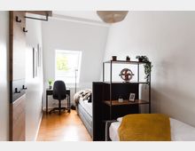 Helles Schlaf- und Arbeitszimmer mit einem Bett, einem Regal und einem Schreibtisch vor einem Dachfenster. Der Raum hat helle Wände, einen Holzboden und ist minimalistisch mit Pflanzen und Dekorationsgegenständen gestaltet.