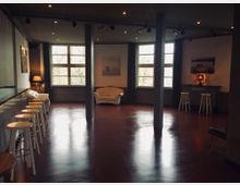 Ein leerer Innenraum mit poliertem Holzboden, großen Fenstern mit Vorhängen und mehreren schwarzen Pfeilern. Der Raum ist eingerichtet mit einer weißen Couch, einer Bar mit Barhockern und Wanddekorationen.