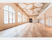Heller, hoher Saal mit großen, bogenförmigen Sprossenfenstern entlang einer Wand und einem Fischgrät-Parkettboden. Die Decke ist reich mit Stuck und geometrischen Kassetten verziert; rechts befindet sich eine moderne Galerieebene mit Glasgeländer und Treppe.