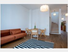 Helles Wohnzimmer mit Holzboden, einer roten Polstercouch, einem runden Holztisch mit drei Stühlen und einem gestreiften Teppich. Im Hintergrund eine Treppe mit Holzstufen, weiße Wände und mehrere Hängeleuchten.