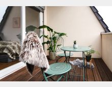 Kleine Balkonfläche mit Holzboden, einem runden, mintgrünen Metalltisch, zwei passenden Stühlen, von denen einer mit einem Fellwurf dekoriert ist. Im Hintergrund stehen große Zimmerpflanzen und weitere Grünpflanzen in Töpfen, angrenzend an eine Glasschiebetür mit Blick ins Innere.