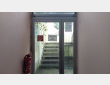 Glastür mit Notausgangsschild und Hinweisen wie „Rauchen verboten“, die zu einer Außentreppe führen; umgeben von leicht verwitterten Wänden mit Pflanzenbewuchs. Links an der Wand ist ein Feuerlöscher montiert.