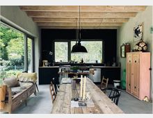 Offener Wohn- und Küchenbereich mit Betonboden, schwarzer Küchenzeile und großer Fensterfront zum grünen Garten. Links führt eine Holztreppe nach oben, im Vordergrund steht ein langer rustikaler Holztisch mit Stühlen und darüber hängt eine schwarze Pendelleuchte.