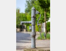 Antiker, verzierter Wasserpumpenbrunnen aus Metall mit gebogenem Griff, stehend auf einem Steinpodest, im Hintergrund eine gepflasterte Straße und grüne Bäume.