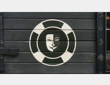 Eine Holzwand in dunklem Grün mit einem aufgemalten Symbol: ein weißer Kreis mit schwarzen Segmenten, der ein stilisiertes, maskenähnliches Gesicht in Schwarz-Weiß zeigt.