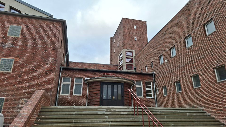Backsteinbau mit moderner Architektur, das den Eingang eines Stadtbads zeigt. Zu sehen sind breite Treppenstufen mit einem roten Geländer, ein zentraler Eingang mit gewölbtem Vordach und hohe, schlichte Fenster.
