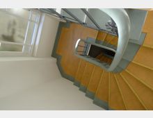 Treppe in einem Gebäude mit gelben Stufen und grauen Kanten, die sich spiralförmig nach unten windet. Links befindet sich ein Fenster mit gefrostetem Glas, das Tageslicht einlässt.