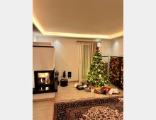 Wohnzimmer mit weihnachtlicher Dekoration: Im Mittelpunkt steht ein geschmückter Weihnachtsbaum mit leuchtenden Lichtern und Geschenken darunter. Links im Bild befindet sich ein Kamin mit brennendem Feuer, daneben stapelweise Kaminholz, während der Boden mit einem orientalischen Teppich bedeckt ist.