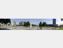 Panoramabild eines städtischen Platzes mit einem zentralen Springbrunnen und umgeben von moderner Architektur, flankiert von Bäumen. Im Vordergrund befinden sich Spazierwege, Sitzbänke und Bereiche für Erholung, während Fahnen vor den Gebäuden wehen.