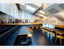 Innenansicht eines schlicht eingerichteten Cafés oder Aufenthaltsraums mit schwarzen Bänken, Holztischen und Holzstühlen. An der Wand hängen auffällige Theaterplakate, und die Decke ist sichtbar mit Holzbalken und Dachfenstern gestaltet.