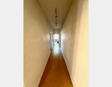 Langer, schmaler Flur mit weißen Wänden und einem braunen Bodenbelag. Am Ende des Flurs ist eine geöffnete Tür zu einem hellen Raum sichtbar, an der Decke eine schlichte Lampenleiste mit mehreren Strahlern.