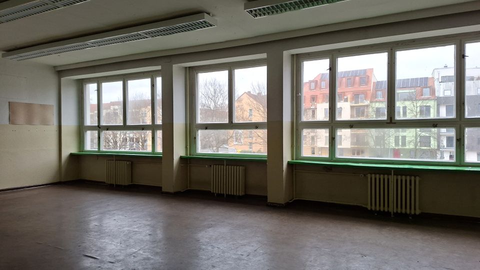 Leerer, älterer Innenraum mit großen Fensterfronten entlang einer Wand, grünen Fensterbänken und Heizkörpern darunter. An der Decke hängen lange Leuchtstofflampen; durch die Fenster sind Wohngebäude und Bäume zu sehen.