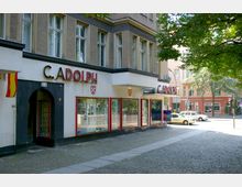 Eckgebäude mit einem Geschäft im Erdgeschoss, das die Aufschrift "C. Adolph" trägt, mit großen Schaufenstern und einem kleinen Eingangsbereich. Der Gehweg besteht aus Kopfsteinpflaster, und im Hintergrund befinden sich weitere farbige Gebäude und Bäume.