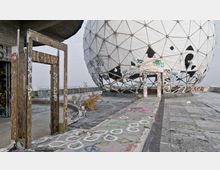 Verlassene Überwachungsstation mit einer geodätischen Kuppel, umgeben von Betonflächen und Wänden, die mit Graffiti bedeckt sind. Im Vordergrund befinden sich ein leeres Metalltürrahmen und verwitterter Boden mit weiteren Graffitimotiven.
