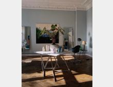 Ein heller Raum mit hohen Decken und dekorativen Stuckverzierungen. In der Mitte steht ein Tisch mit Pflegeprodukten und einer großen Zimmerpflanze, im Hintergrund sind ein Gemälde, ein Spiegel und eine moderne Sitzgruppe sichtbar.