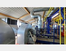 Innenraum einer Industrieanlage mit großen Metallrohren, Maschinen und gelben sowie blauen Rohrleitungen. Das Gebäude hat eine gewölbte Decke aus Metall und Holzträgern.