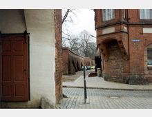 Straßenszene in einer historischen Stadt mit roten Backsteingebäuden, darunter ein Gebäude mit Erker und ein gepflasterter Gehweg. Im Hintergrund sind eine Backsteinmauer mit Stützpfeilern und karge Bäume sichtbar.