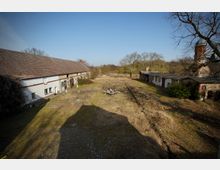 Weitläufiger, verwilderter Gutshofhof mit gras- und brachbewachsenem Gelände zwischen langen, niedrigen Wirtschaftsgebäuden mit Ziegeldächern. Im Hintergrund stehen kahle Bäume, rechts ist ein runder Backsteinturm mit Kuppeldach am Rand der Anlage zu sehen.