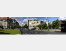 Städtische Straßenszene mit mehrspuriger Fahrbahn, flankiert von Bäumen, Grünflächen und Gebäuden, darunter ein historisches Haus mit rotem Dach und ein moderner Gebäudekomplex mit der Aufschrift "Deutsche Bank". Im Vordergrund stehen Laternen und eine Haltestelle, rechts ist ein Teil eines Doppeldeckerbusses sichtbar.