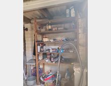 Innenansicht eines kleinen Abstellraums oder einer Werkstatt mit weißen Ziegelwänden und einem großen Holzregal voller Werkzeuge, Reinigungsmittel und verschiedener Gegenstände. Im Vordergrund stehen ein Staubsaugerschlauch und weitere gelagerte Utensilien, darüber verläuft eine Deckenleuchte unter einer Holzbalkendecke.