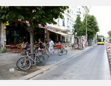 Straßenszene in einer Stadt mit Cafés und Geschäften entlang eines Gehwegs, mehreren geparkten Fahrrädern, Bäumen und einem vorbeifahrenden gelben Linienbus auf einer schmalen Straße. Einige Menschen sitzen an Tischen im Freien, während andere vorbeigehen.