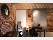 Industriell gestalteter Raum mit Ziegelsteinwänden, einer Holzkommode, einem rechteckigen Spiegel, einem Schreibtisch mit Lampe sowie einem Stuhl mit Fellbezug. Die Decke zeigt gemusterte Ziegel und ein modernes Metallpaneel ziert die Wand.