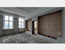 Ein leerer Raum in einem renovierungsbedürftigen Zustand mit großen Fenstern, die viel Tageslicht einlassen. Die Wände sind teilweise unverputzt, und der Boden zeigt Abnutzungsspuren; eine offene Wandöffnung führt in einen weiteren Raum.