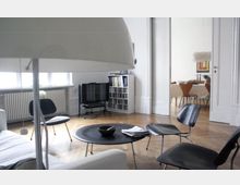 Wohnzimmer mit Holzboden, modernen schwarzen Stühlen und einem runden Couchtisch. Im Hintergrund sind ein kleiner Fernseher, ein Regal mit CDs und durch eine offene Tür ein Essbereich mit Holztisch und Stühlen sichtbar.