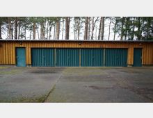 Ein langes Holzgebäude mit fünf grünen Garagentoren aus Metall und zwei Seitentüren, umgeben von einem Betonvorplatz und hohen Nadelbäumen im Hintergrund.