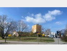 Städtische Szenerie mit einem mehrstöckigen gelben Gebäude im Zentrum, umgeben von kahlen Bäumen und blühenden gelben Sträuchern auf einer Grünfläche. Im Vordergrund ein Gehweg, eine Straße und eine Fußgängerampel, im Hintergrund sind weitere Gebäude sichtbar.