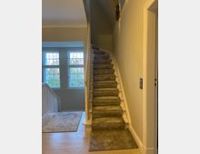 Eine schmale, nach oben führende Treppe mit grauem Teppich in einem hell gestrichenen Innenraum. Links befindet sich ein Fenster mit weißem Rahmen und Blick auf Bäume, daneben ein Treppenabsatz mit hellem Holzfußboden und einem Läufer.