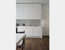 Moderne Küche mit weißen, glatten Einbauschränken und weißen Arbeitsflächen. Der Boden ist aus dunklem Holz, die Wand besitzt eine hellgraue Fliesenrückwand, und sichtbar sind ein Wasserhahn, eine Spüle sowie ein Esstisch mit Holzstuhl im Vordergrund.