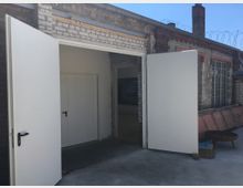 Außenansicht eines Gebäudes mit einer offenen weißen Doppeltür, die in einen Innenraum führt. Die Umgebung zeigt helles Mauerwerk, ein Glasfenster rechts sowie einen gepflasterten Boden und eine einfache metallene Feuerschale im Vordergrund.