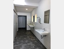 Eine moderne, helle öffentliche Waschräumeinrichtung mit grauen Fliesen, weißen Wänden und mehreren Waschbecken auf der rechten Seite, ergänzt durch spiegelartige Wandflächen. Links sind geschlossene Kabinentüren zu sehen, und im Hintergrund befindet sich eine Tür mit Metalloptik.