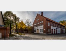 Straßenszene mit einem zweistöckigen roten Backsteingebäude, das die Aufschrift „Turnhalle - Feuerwehr“ und „Freiwillige Feuerwehr“ trägt, sowie einer gepflasterten Straße und Bäumen mit herbstlichem Laub. Im Hintergrund sind weitere Backsteingebäude und parkende Autos zu sehen.