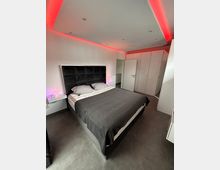 Modernes Schlafzimmer mit einem Doppelbett in dunkler Bettwäsche und gesteppter Rückenlehne, flankiert von kleinen Nachttischen. Im Hintergrund stehen weiße Einbauschränke, und die Decke ist mit roten LED-Leuchten indirekt beleuchtet.