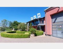 Ein moderner Gebäudekomplex mit rosafarbener Fassade und großen Glasfenstern, über dem die Buchstaben "TSH" auf Schildern angebracht sind. Im Vordergrund befindet sich ein gepflegter Garten mit symmetrischen Buchsbaumhecken und dekorativen Pflanzen.