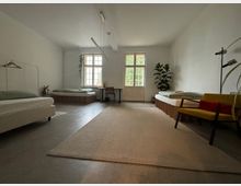 Ein schlicht eingerichtetes Zimmer mit drei Betten an den Wänden, einem großen Fenster mit Doppeltür in der Mitte, einem Teppichboden und minimalistischer Dekoration wie Pflanzen, einer Stehlampe und einem gelben Sessel. Die Wände sind weiß, und der Boden besteht aus dunklem Beton.