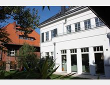 Zwei benachbarte Gebäude, eins mit weißer Fassade und großen Fenstern im modernen Stil, das andere mit roten Ziegeln und einem Dach aus roten Ziegeln. Im Vordergrund befindet sich ein Garten mit Rasen, Bäumen und einem Holzgitter.