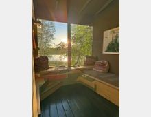 Gemütlicher Innenraum mit Holzboden und einer Eckbank, die mit Kissen und Decken dekoriert ist, durch ein großes Fenster mit Blick auf eine baumreiche Landschaft und einen See bei Sonnenuntergang.