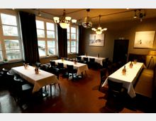 Innenraum eines Restaurants mit mehreren langen, weiß gedeckten Tischen und schwarzen Stühlen, beleuchtet von Deckenleuchten und einer Spiegelkugel. Große Fenster mit dunklen Vorhängen zeigen einen Blick auf Wasser und eine Brücke; an den grauen Wänden hängen gerahmte Bilder.